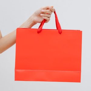 Crop unrecognizable female showing red paper shopping bag in hand against white wall
